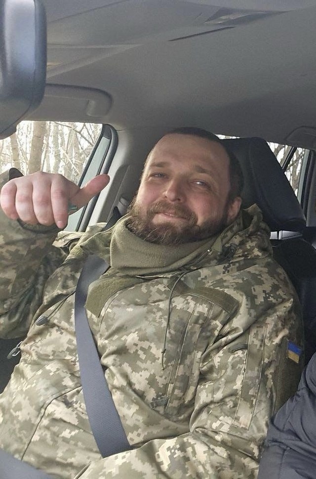 Pavlo Lukin in uniform in the passenger seat of a car, smiling, giving a thumbs up.